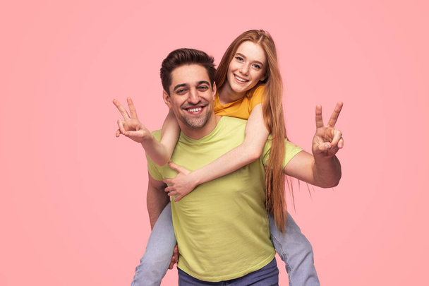 Optimistic young man and woman in colorful clothes gesturing V sign and looking at camera with smile against pink background - Foto, Imagen
