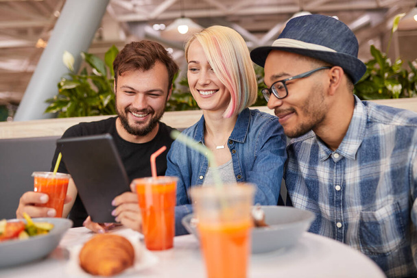 Boldog változatos férfiak és nők gyümölcslé mosolygós és videó nézése tabletta, miközben ül az asztalnál, miközben a barátok találkoznak a modern étteremben - Fotó, kép