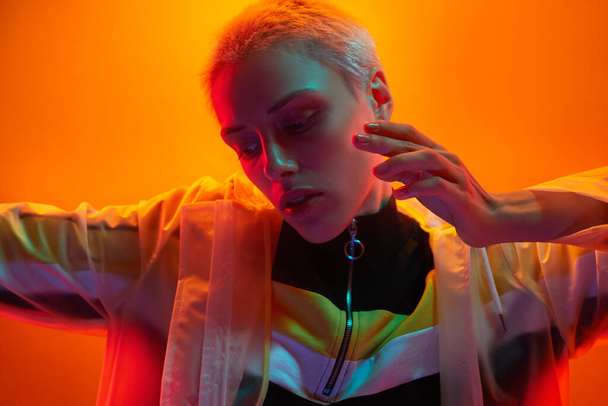 Young androgynous woman in futuristic outfit stretching out arm and looking down while touching face under neon illumination against orange background - Photo, image