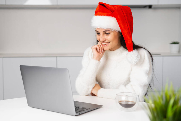 Μια γυναίκα με κόκκινο Αϊ Βασίλη παρακολουθεί μια ταινία ή βίντεο και χαμογελά χρησιμοποιώντας το laptop της κατά τη διάρκεια των Χριστουγέννων.  - Φωτογραφία, εικόνα