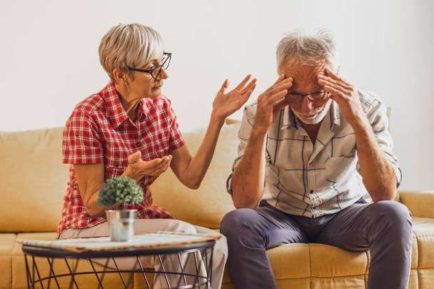 Senior couple sitting in living room. Man is sad and worried and woman is consoling him. - Photo, image