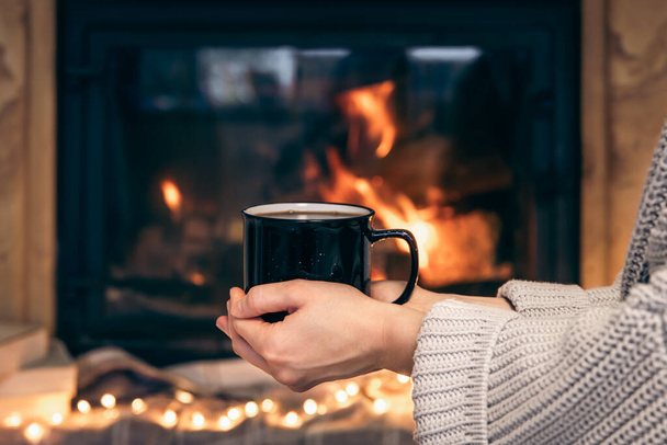Ένα φλιτζάνι με ένα ζεστό ρόφημα σε γυναικεία χέρια σε ένα θολό φόντο ενός τζακιού, ένα ζεστό φόντο Χριστουγέννων. - Φωτογραφία, εικόνα