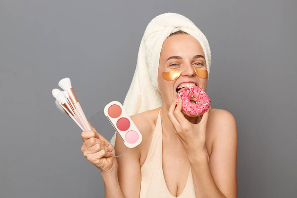 Portrait of excited hungry smiling woman wearing top and towel on her head isolated over gray background, holding brushes and eyeshadow, biting delicious donut. - Foto, afbeelding
