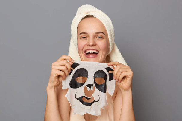 Photo of smiling happy delighted woman wearing top and towel on her head isolated over gray background, showing panda facial mask, laughing happily, doing skin care procedures. - Fotografie, Obrázek
