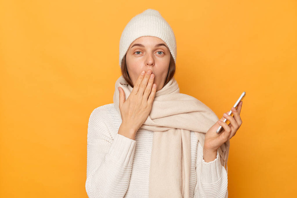 Wizerunek zaszokowanej brązowłosej młodej dorosłej kobiety ubranej w ciepły sweter, czapkę i szalik, używającej telefonu komórkowego, zakrywającej usta dłonią, czytającej coś zaskakującego, pozującej na żółtym tle. - Zdjęcie, obraz