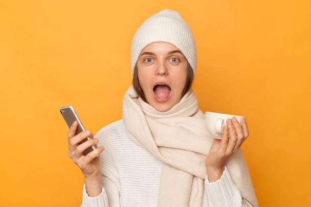 Image of amazed surprised Caucasian woman wearing warm jumper, cap and scarf posing isolated over yellow background, keeps mouth widely open, holding mobile phone in hands. - Photo, image