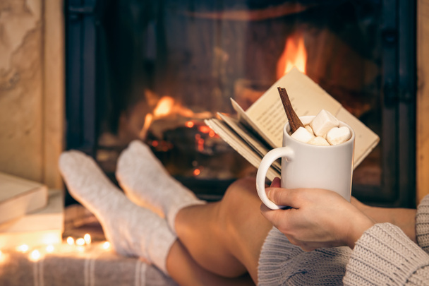 Женщина держит чашку какао с зефиром и книгу, сидя у камина дома. - Фото, изображение