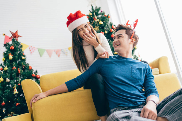 サンタクロースの帽子をかぶっている若い美しい幸せなアジアの女性は驚きと背景にクリスマスツリーで自宅で彼女のボーイフレンドの後ろに笑う。コピースペース付き画像. - 写真・画像