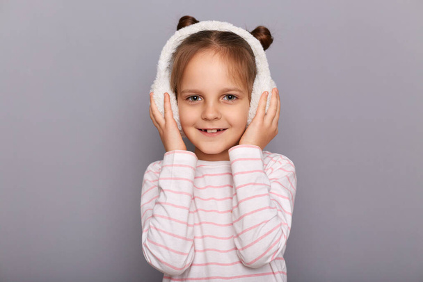Portrait of little cute female kid standing in fur headphones and looking at camera with smile, expressing positive emotions, wearing striped shirt, being in good mood. - Photo, Image