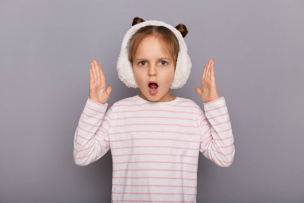 Portrait of shocked astonished little girl wearing striped shirt and fur headphones standing isolated over gray background, raised her arms, sees something shocked, keeps mouth opened. - Photo, Image