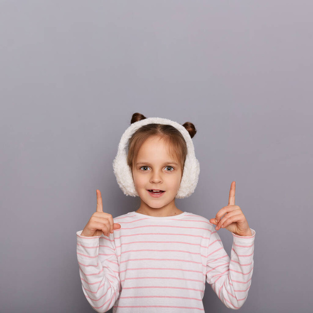 Indoor shot of amazed little girl wearing striped shirt and fur headphones standing isolated over gray background, pointing index fingers up at advertisement area, copy space for promotion. - Foto, immagini