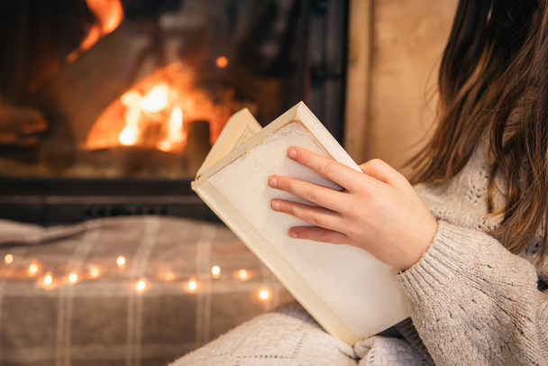Primer plano, una niña lee un libro sobre un fondo borroso de una chimenea en llamas. - Foto, imagen