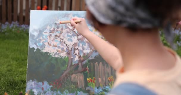 Közelkép egy női művész kezéről, amint vásznat rajzol egy zöld mezőn tavasszal. Festőműhely a kertben. A művészeti oktatás fogalma. Szabadtéri szabadidős tevékenységek - Felvétel, videó