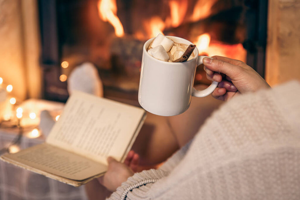 Una donna tiene una tazza di cacao con marshmallow e un libro mentre è seduta accanto al camino a casa. - Foto, immagini