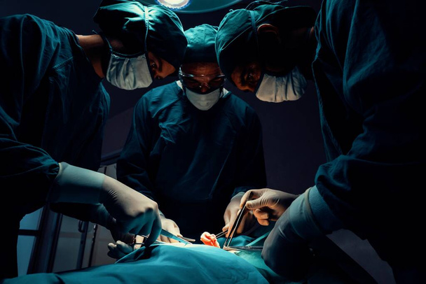 Χειρουργική ομάδα εκτελεί χειρουργική επέμβαση σε ασθενή σε αποστειρωμένο χειρουργείο. Σε ένα χειρουργείο που φωτίζεται από μια λάμπα, μια επαγγελματική και σίγουρη χειρουργική ομάδα παρέχει ιατρική φροντίδα σε έναν αναίσθητο ασθενή.. - Φωτογραφία, εικόνα