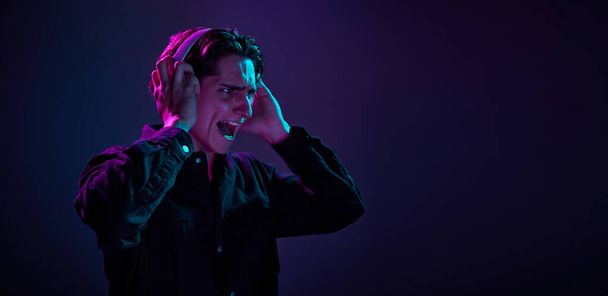 Portrait of young man in casual black shirt isolated on gradient dark purple background in neon light. Music in headphones. Concept of human emotions, facial expression, sales, ad, fashion and beauty - Фото, зображення
