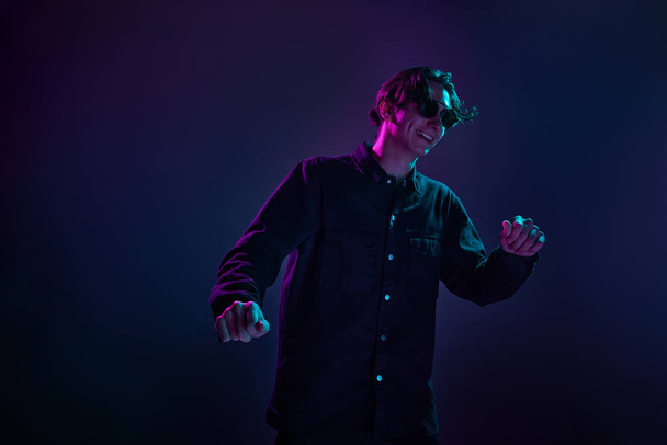 Portrait of young man in casual black shirt posing in sunglasses isolated over gradient dark purple background in neon light. Smile. Concept of human emotions, facial expression, sales, ad, fashion - Valokuva, kuva