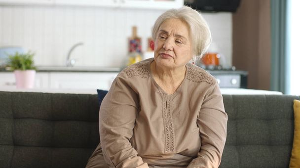 自宅や老人ホームで思慮深い老婦人。高齢者のための保健サービス。彼女の左に空白の広告スペースを見てリビングルームでソファに座っている老婦人の肖像画. - 写真・画像