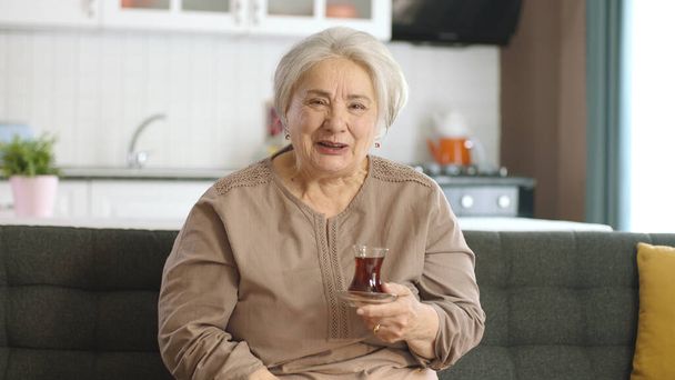 Щаслива стара жінка дивиться цікавий фільм по телевізору під час пиття чаю. Стара жінка сидить на дивані, дивлячись таємничий телефільм сам. Старі люди. Почуття
. - Фото, зображення