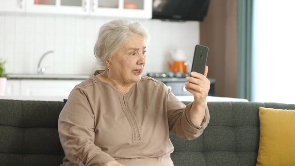 Oudere vrouw maakt videogesprek met familieleden, kijkt naar de camera en zwaait, zit op een comfortabele bank. Technologiegebruiksconcept met senioren. - Foto, afbeelding