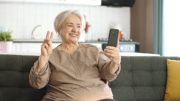 Mujer mayor sonriendo, haciendo videollamadas con familiares, mirando a la cámara y saludando, sentada en un cómodo sofá. La tecnología utiliza el concepto con los ancianos. Tomarse una selfie con una señal de victoria. - Foto, imagen