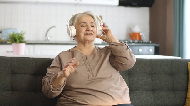 Colorful and funny moments of an old woman listening to music with headphones on her sofa. Funny old woman with headphones singing and dancing. Mature cheerful woman enjoying life after retirement. - Zdjęcie, obraz
