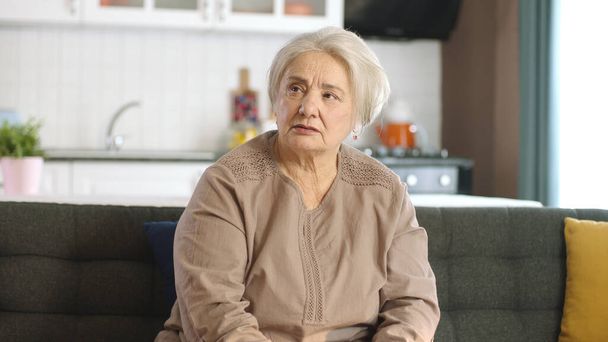 Smutna staruszka w domu lub w domu opieki. Usługi zdrowotne dla osób starszych. Portret staruszki siedzącej na kanapie w salonie, patrzącej na pustą przestrzeń reklamową po prawej stronie ekranu. - Zdjęcie, obraz