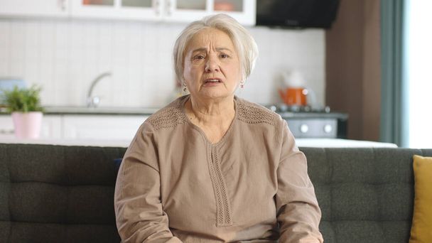 Nachdenkliche alte Frau in ihrem Heim oder Pflegeheim. Gesundheitsdienste für ältere Menschen. Porträt einer älteren Frau, die auf dem Sofa im Wohnzimmer in die Kamera blickt. - Foto, Bild