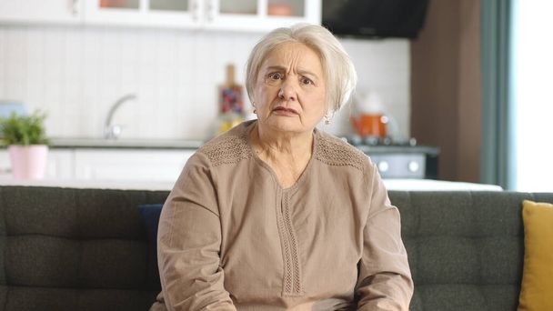 Στοχαστική ηλικιωμένη γυναίκα στο σπίτι της ή στο γηροκομείο. Υπηρεσίες υγείας για ηλικιωμένους. Πορτρέτο της ηλικιωμένης γυναίκας κοιτάζοντας κάμερα, ενώ κάθεται στον καναπέ στο σαλόνι. - Φωτογραφία, εικόνα