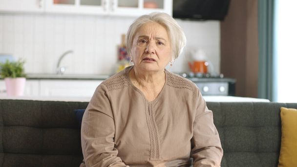 Eine ältere Frau mit weißem Haar streitet sich bei einem Online-Videoanruf mit der anderen Person. Wütend beunruhigte Seniorin telefoniert auf Laptop auf Sofa im Wohnzimmer zu Hause. Ansicht der Frontkamera. - Foto, Bild