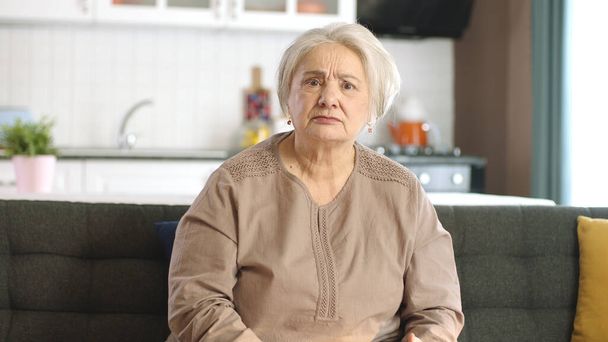 Una anciana pensativa en su casa o en su residencia. Servicios de salud para ancianos. Retrato de una anciana mirando a la cámara mientras está sentada en el sofá en la sala de estar. - Foto, imagen