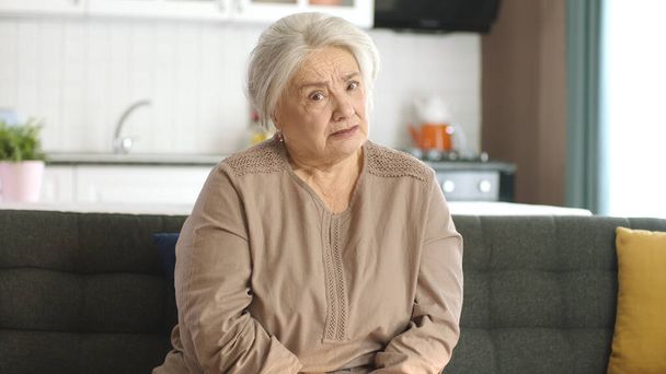 Una anciana pensativa en su casa o en su residencia. Servicios de salud para ancianos. Retrato de una anciana mirando a la cámara mientras está sentada en el sofá en la sala de estar. - Foto, imagen