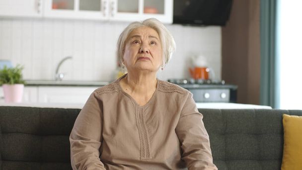 Unhappy and grumpy old woman is resting in her quiet home. Lonely old woman sitting alone on the sofa looking at the empty advertising space above the screen. - Photo, Image