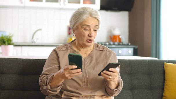 La anciana mira sus cuentas de redes sociales, lee las noticias, escribe mensajes en sus dos teléfonos inteligentes. Una anciana que intenta usar dos smartphones se sorprende por lo que ve en la pantalla. - Foto, imagen