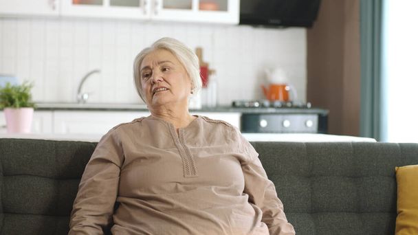 Huomaavainen vanha nainen kotonaan tai hoitokodissaan. Terveyspalvelut vanhuksille. Muotokuva vanha nainen istuu sohvalla olohuoneessa katsoen tyhjä mainostilaa hänen vasemmalla. - Valokuva, kuva