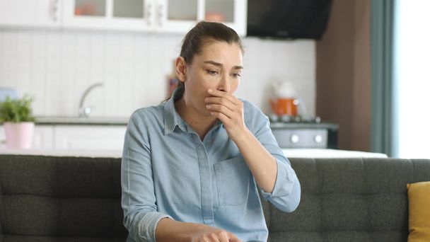 A young woman who is sick is coughing. A young woman in her 30s having difficulty breathing in the living room of her house. Shortness of breath, asthma, trouble breathing. Corona Virus symptoms. - Photo, Image