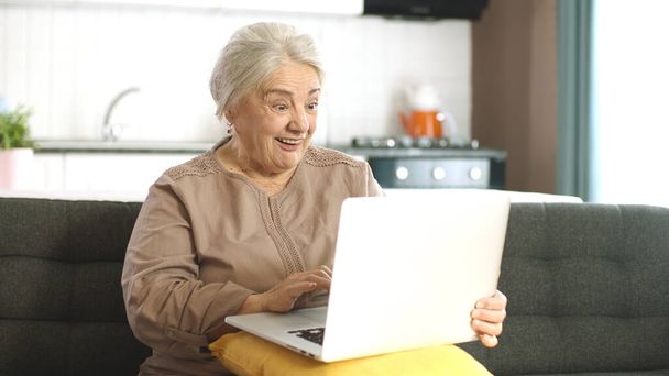 Donna anziana matura trascorrere del tempo navigando in internet sul computer portatile. Una donna sulla settantina sta facendo una videochiamata in chat virtuale. Senior adulto signora shopping online sul divano in soggiorno. - Foto, immagini
