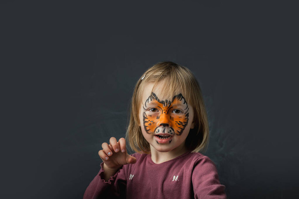 Nettes kleines kaukasisches Mädchen mit Tiger-Gesichtsbemalung auf schwarzem Hintergrund. Nahaufnahme Porträt eines kleinen Kindes mit Kinderschminken. Jahr eines Tigers. Glücklich lächelndes Mädchen, das vorgibt, ein Tiger zu sein - Foto, Bild