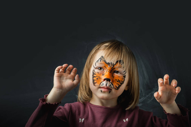 Schattig klein blank meisje met tijger gezicht schilderen op de zwarte achtergrond. Close-up portret van kleine jongen met gezicht-schilderij. Jaar van een tijger. Gelukkig lachend meisje dat doet alsof ze een tijger is - Foto, afbeelding