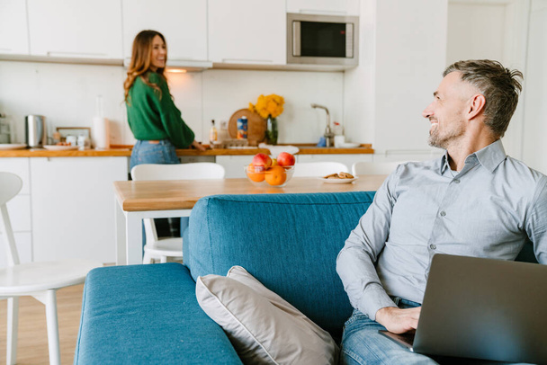 marido feliz con el ordenador portátil mirando a su esposa en la cocina en casa - Foto, imagen