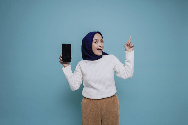Excited Asian Muslim woman wearing white shirt and hijab pointing at the copy space on top of her and showing phone screen, isolated by blue background - Photo, image