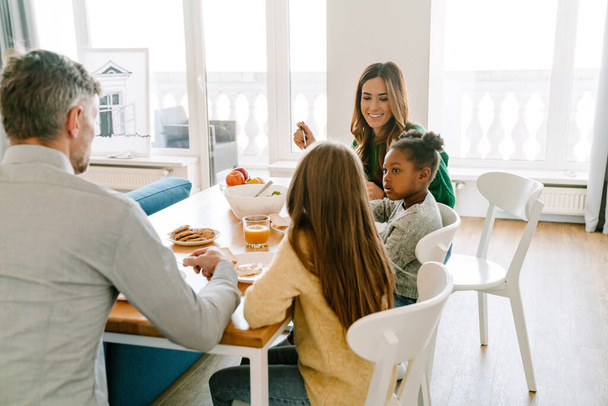 Happy family with adopted afro girl eating breakfast together in cozy kitchen at home. Adoption concept. - Фото, изображение