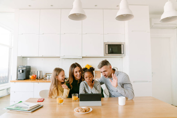 Счастливая улыбающаяся семья, разговаривающая по видеосвязи с родственниками, сидя дома за кухонным столом - Фото, изображение