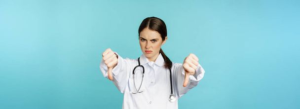 Woman doctor, medical worker showing thumbs down and frowning angry, dislike, disappove smth, standing in white coat over blue background. - 写真・画像