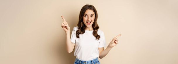 Happy authentic girl smiling, pointing fingers sideways, showing left and right banner, demonstrating promo, standing against beige background. - Photo, Image