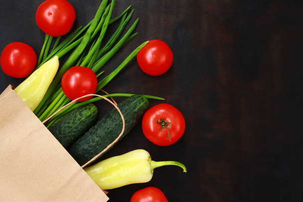 Paper bag with spilled vegetables on a dark wooden table, top view. Ripe tomatoes, green onions, cucumbers and yellow peppers on the table. The concept of healthy eating and vegetarianism. Buying vegetables - Photo, Image