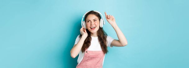 Νεαρή γυναίκα χορεύει και ακούει μουσική με ακουστικά, σηκώνει το χέρι και χαμογελά ανέμελα, απολαμβάνοντας το αγαπημένο τραγούδι, στέκεται πάνω από το μπλε φόντο. - Φωτογραφία, εικόνα