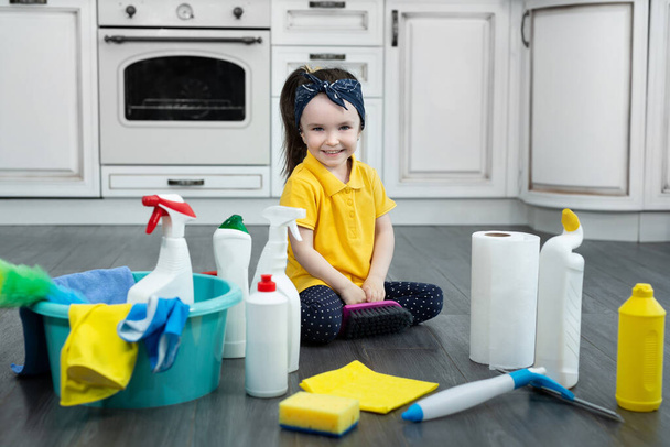 Niña juega con detergentes y un cepillo en el suelo de la cocina mientras limpia - Foto, Imagen