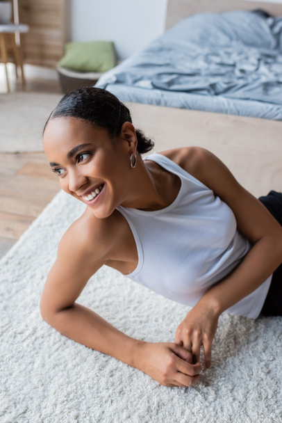 young and joyful african american woman in tank top sitting on carpet  - Foto, afbeelding