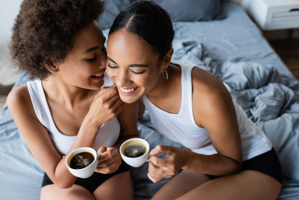 high angle view of happy lesbian african american couple holding cups of coffee while sitting on bed - Photo, Image